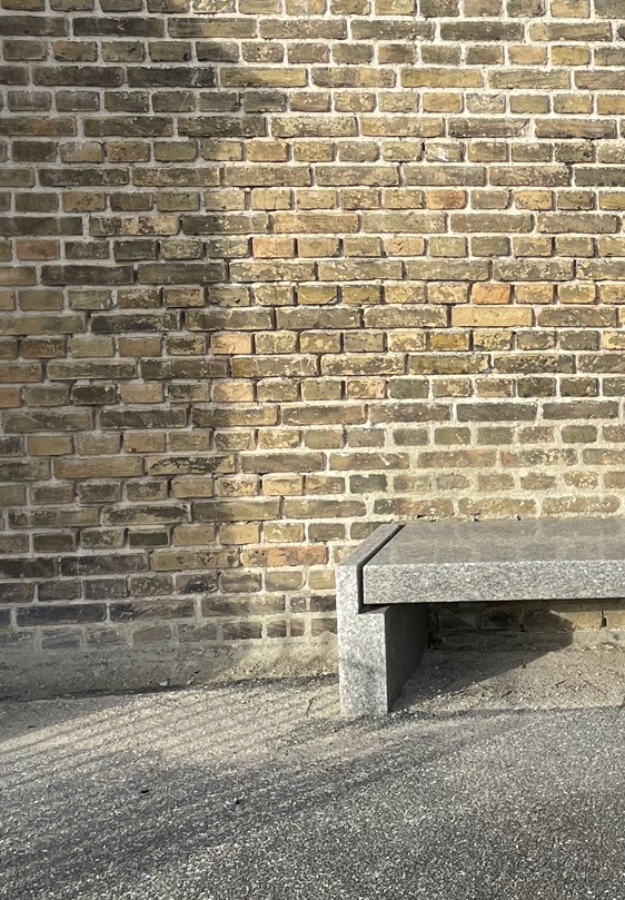 Enden af stenbænk foran en gul murstensvæg, med efterårets sol og skygger.